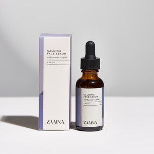 May include: A brown glass bottle with a black dropper top filled with a clear liquid. The bottle is labeled "CALMING FACE SERUM with Lavender +  Jojoba 1 FL OZ" and "ZAAINA". The bottle is next to a white box with the same label.