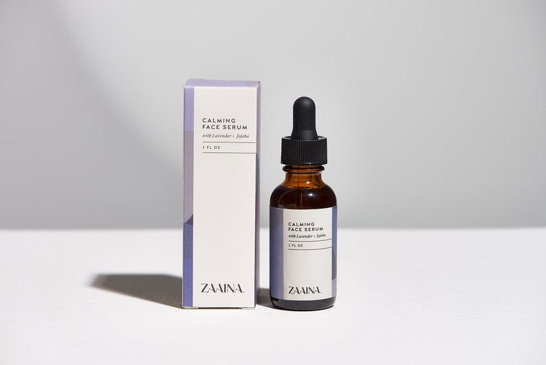 May include: A brown glass bottle with a black dropper top filled with a clear liquid. The bottle is labeled "CALMING FACE SERUM with Lavender +  Jojoba 1 FL OZ" and "ZAAINA". The bottle is next to a white box with the same label.