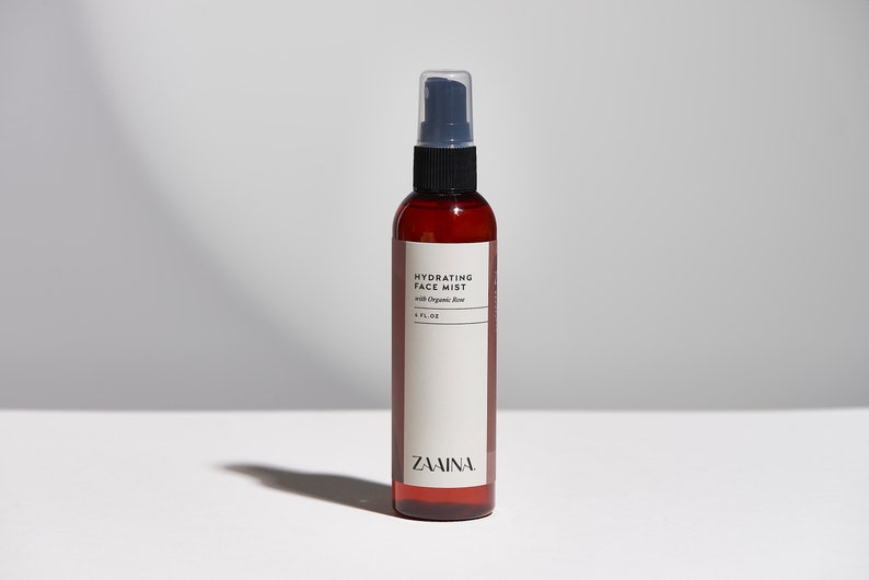Puede incluir: Una botella de vidrio marr&oacute;n con una boquilla de pulverizaci&oacute;n negra y una etiqueta blanca que dice "Hydrating Face Mist with Organic Rose 4 FL OZ ZAINA."
