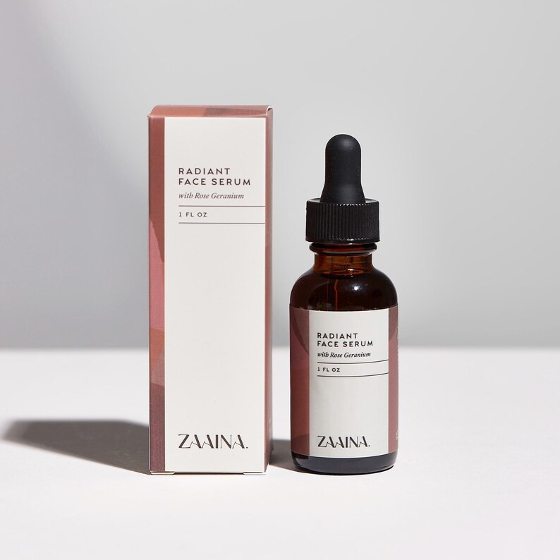 K&ouml;nnte beinhalten: Eine 30 ml Braunglasflasche mit Radiant Face Serum with Rose Geranium und schwarzem Pipettenverschluss, neben der passenden rechteckigen Pappschachtel. Beide tragen wei&szlig;e Etiketten mit schwarzer Schrift und dem Markennamen Zaaina.
