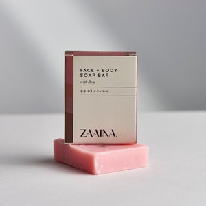 Puede incluir: Una pastilla de jab&oacute;n rosa con el texto "FACE + BODY SOAP BAR with Rose 1.2 OZ / 34 GM ZAMINA." impreso en el embalaje.