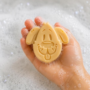 May include: A light tan, solid bar of soap shaped like a cartoon dog head with floppy ears, a rounded snout, and a small tongue sticking out. The soap has a smooth, molded surface and is covered in white soap bubbles from being used in water.