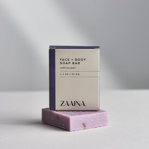 May include: A white box with purple trim and the text "FACE + BODY SOAP BAR with Lavender 1.1 OZ / 31 GM ZAWINA." A purple soap bar with dried lavender flowers is on top of the box.