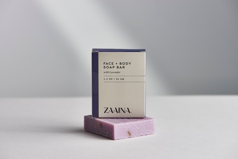 May include: A white box with purple trim and the text "FACE + BODY SOAP BAR with Lavender 1.1 OZ / 31 GM ZAWINA." A purple soap bar with dried lavender flowers is on top of the box.