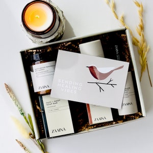 May include: A gift box filled with self-care items, including a soy candle, a face and body soap bar, a moisturizing balm, and a card that reads "Sending Healing Vibes".