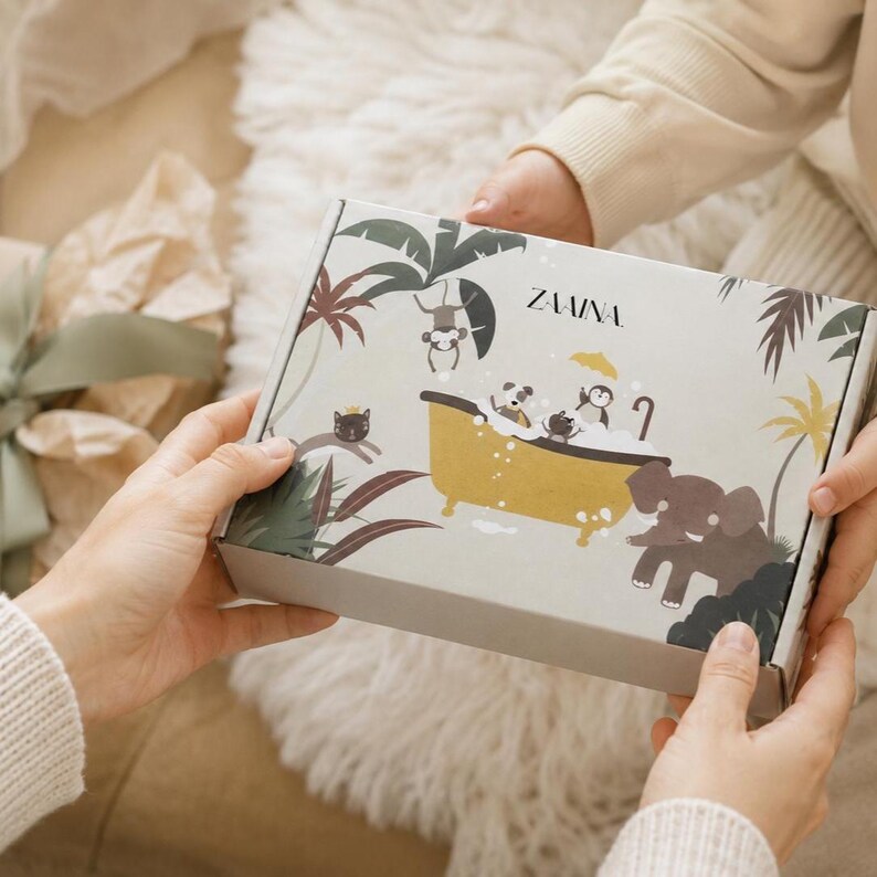 May include: A rectangular cardboard box with a light gray background featuring a colorful jungle-themed illustration. The design includes a yellow bathtub filled with bubbles, various animals, palm trees, and the text ZAAINA printed in black.