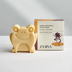 May include: A light beige frog-shaped soap bar next to its product box. The box is labeled KIDS NATURAL SOAP BAR with Shea + Cocoa Butter, FROG SHAPED SOAP, and the brand name ZAAINA, featuring a graphic of a purple frog on a lily pad.