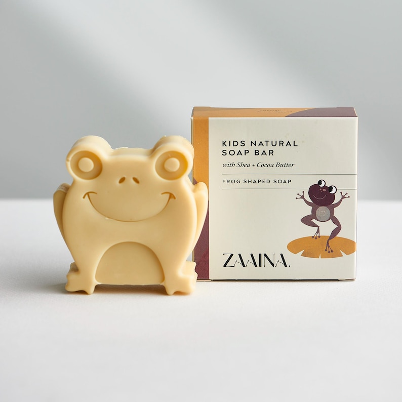 May include: A light beige frog-shaped soap bar next to its product box. The box is labeled KIDS NATURAL SOAP BAR with Shea + Cocoa Butter, FROG SHAPED SOAP, and the brand name ZAAINA, featuring a graphic of a purple frog on a lily pad.