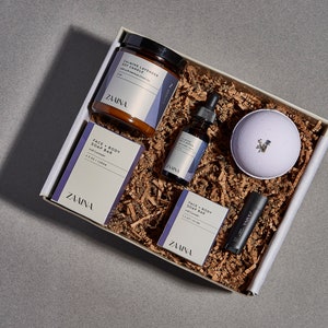 May include: A gift box filled with lavender-themed bath and body products. The box contains a candle, bath bomb, soap bar, lip balm, and essential oil. The products are all labeled with the brand name "Zamina".