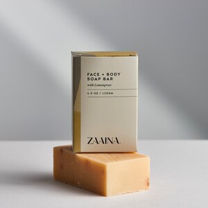 K&ouml;nnte beinhalten: Eine wei&szlig;e Box mit goldenen Akzenten und dem Text "FACE + BODY SOAP BAR with Lemongrass 4.5 OZ / 128GM ZAINA." Die Box liegt auf einer Seife.