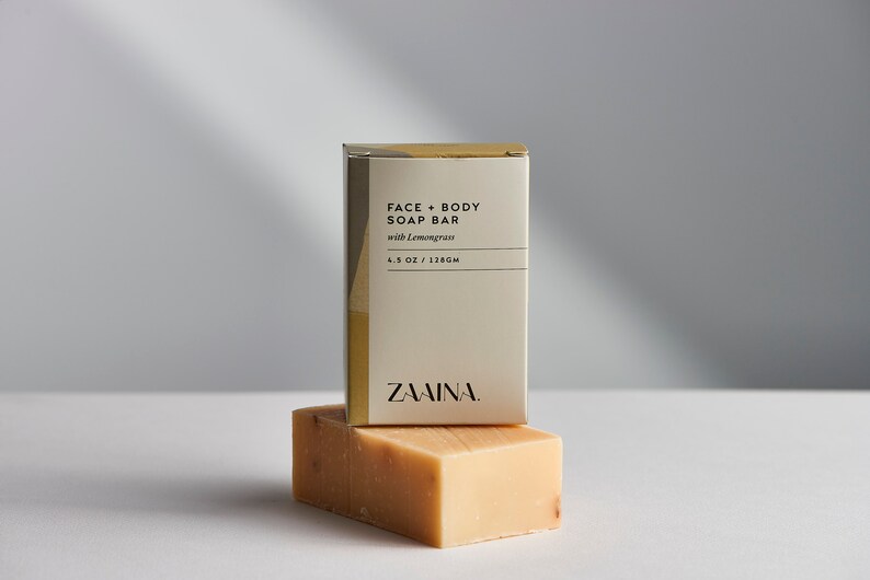 K&ouml;nnte beinhalten: Eine wei&szlig;e Box mit goldenen Akzenten und dem Text "FACE + BODY SOAP BAR with Lemongrass 4.5 OZ / 128GM ZAINA." Die Box liegt auf einer Seife.