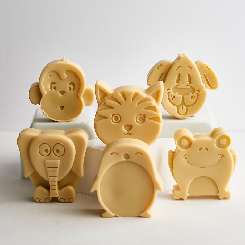 May include: A collection of six light beige animal-shaped soaps featuring molded designs of a monkey, a cat, a dog, an elephant, a penguin, and a frog. Each soap has a smooth, matte finish with detailed facial features embossed on the front surface.