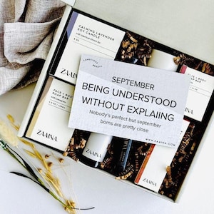 May include: An open gift box filled with beauty products, including a soy candle, soap bar, and lip balm. A card reads "September, Being Understood Without Explaining." The box contains decorative filler and is presented on a white surface.