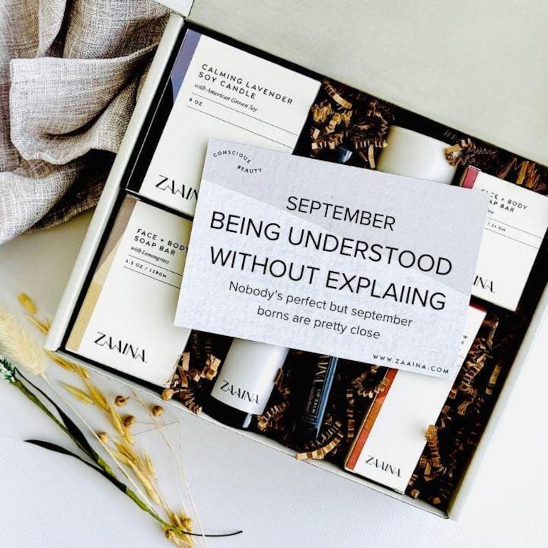 May include: An open gift box filled with beauty products, including a soy candle, soap bar, and lip balm. A card reads "September, Being Understood Without Explaining." The box contains decorative filler and is presented on a white surface.