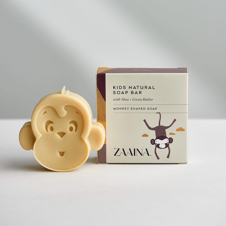 May include: A light beige monkey-shaped soap bar next to its square cardboard packaging. The box features the text KIDS NATURAL SOAP BAR with Shea + Cocoa Butter, MONKEY SHAPED SOAP, and ZAAINA, along with a graphic illustration of a hanging monkey.