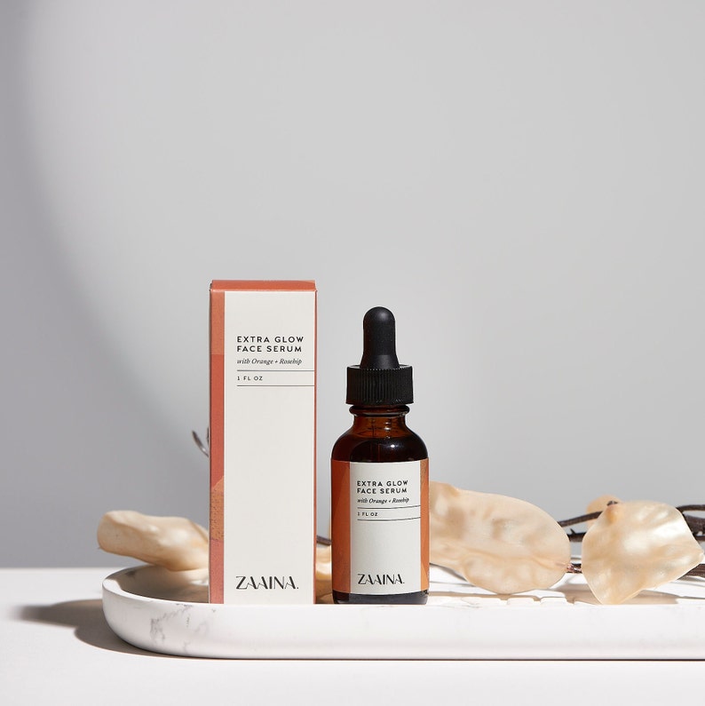 May include: A brown glass bottle with a black dropper top containing a face serum. The bottle is labeled "Extra Glow Face Serum with Orange + Rosehip" and "ZAINA". The bottle is sitting on a white marble tray with a light brown branch behind it. The bottle is next to a white box with the same label.
