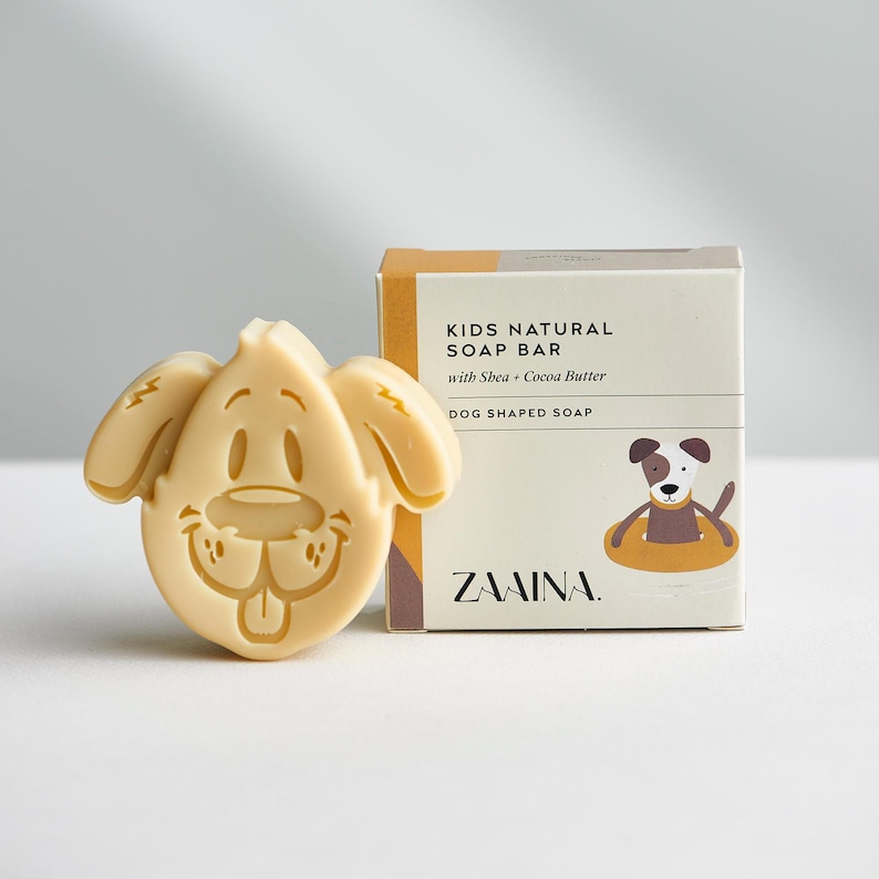 May include: A light beige dog-shaped soap bar next to its square cardboard packaging. The box features the text KIDS NATURAL SOAP BAR with Shea + Cocoa Butter, DOG SHAPED SOAP, and ZAAINA, along with a graphic of a dog in a yellow floatie.