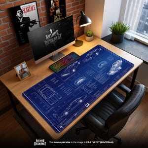 Puede incluir: Una alfombrilla de escritorio azul con planos de coches blancos y el texto "ARTHUR DESKMAT". La alfombrilla mide 60 cm x 120 cm. Un ordenador, un teclado y una lámpara de escritorio están sobre un escritorio de madera.