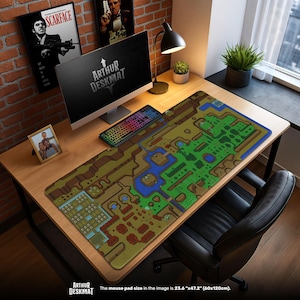 May include: A large desk mat featuring a colorful map design, with shades of green, brown, and blue. The mat is rectangular and covers a wooden desk, with a computer, keyboard, and other accessories. The text "ARTHUR DESKMAT" is visible.