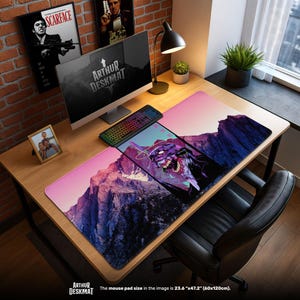 Pode incluir: Um tapete de mesa grande com um design de paisagem montanhosa em tons de rosa, roxo e azul. O tapete apresenta um gráfico de um monstro e o texto "ARTHUR DESKMAT". O tamanho do tapete do rato é 60 cm x 120 cm.