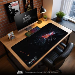 May include: A black desk mat featuring a graphic design from The Witcher III: Wild Hunt. The mat, measuring 23.6" x 47.2", has a central design with red and white accents. The text "ARTHUR DESKMAT" is at the bottom left.