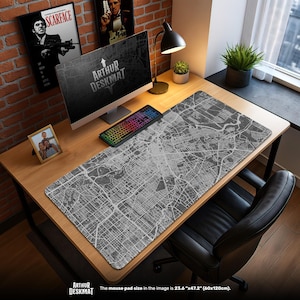 Può includere: Un grande tappetino da scrivania grigio con un design dettagliato di mappa della città, 60 cm x 120 cm. Il tappetino è su una scrivania di legno con un computer, tastiera e lampada. Il testo "ARTHUR DESKMAT" è visibile.