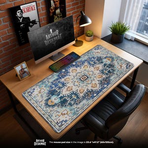 Op de afbeelding: Een rechthoekige bureaumat met een blauw, beige en wit bloemenpatroon, dat op een tapijt lijkt. De mat, met een afmeting van 60cm x 120cm, ligt op een houten bureau met een computer, toetsenbord en lamp. De tekst "ARTHUR DESKMAT" is zichtbaar.