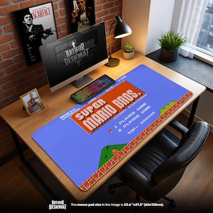 May include: A blue desk mat featuring the Super Mario Bros. game design. The mat has a pixelated landscape with the game's logo and text. The desk mat measures 23.6" x 47.2". A computer, keyboard, and other items are on a wooden desk.