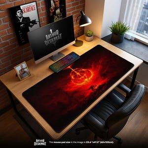 Könnte beinhalten: Eine große, schwarze Schreibtischunterlage mit einem feurigen rot-orangen Grafikdesign. Die Matte zeigt eine Silhouette und ein leuchtendes kreisförmiges Muster. Der Text "ARTHUR DESKMAT" ist sichtbar. Größe: 60cm x 120cm.