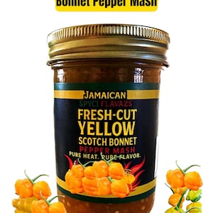 May include: A jar of Jamaican Authentic Yellow Scotch Bonnet Pepper Mash. The jar has a gold lid and a black label with yellow text that reads "FRESH-CUT YELLOW SCOTCH BONNET PEPPER MASH". Several yellow scotch bonnet peppers are arranged around the jar.