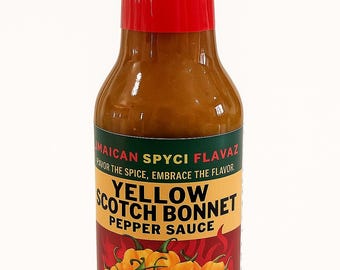 Op de afbeelding: Een fles Yellow Scotch Bonnet Pepper Sauce met een rode dop en een etiket met gele pepers. Op het etiket staat "Jamaican Spyci Flavaz" en "Hot & Spicy". De fles bevat 148 ml.