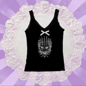 May include: A black sleeveless vest top featuring a white lace trim along the V-neckline and a small white bow centred below the collar. The front of the garment displays a white graphic print of a grid-patterned face with protruding spikes.