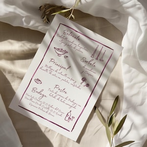 Puede incluir: Un menú impreso con texto manuscrito e ilustraciones en tinta morada oscura. El menú incluye secciones para Entrante, Principal, Postre y Bodega. El papel está sobre un fondo de tela blanca con ramitas de vegetación.