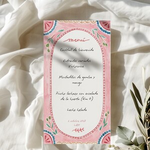 Op de afbeelding: Een aquarel menukaart met een roze rand en decoratieve bloemelementen. Het menu, in het Spaans, bevat gerechten zoals "Cocktail de bienvenida" en "Pincho lechazo con ensalada". De kaart ligt op een witte stoffen achtergrond.