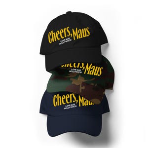 Könnte beinhalten: Ein Stapel Baseballkappen in Schwarz, Camouflage und Marineblau. Jede Kappe trägt den gelben Schriftzug "Cheers, Maus" mit "Love Aus Hollywood" darunter. Die Kappen sind auf weißem Hintergrund gestapelt.
