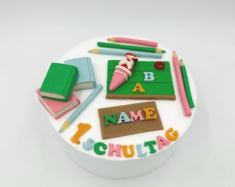 May include: A round cake decorated with school-themed fondant figures. The cake features miniature books, pencils, a chalkboard with letters, and the words "NAME" and "1. SCHULTAG" in colorful letters.