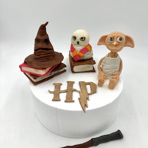 Könnte beinhalten: Kuchendekorationen mit Harry-Potter-Motiv. Das Set enthält einen braunen Sprechenden Hut auf Büchern, eine weiße Eule mit Schal, eine Dobby-Figur, einen Zauberstab und die Buchstaben "HP" mit einem Blitz.