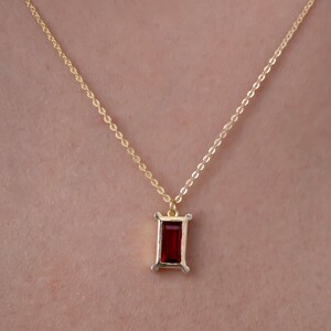 May include: A gold-colored necklace with a rectangular, deep red gemstone pendant. The pendant is framed by a gold-colored setting. The necklace chain is delicate and visible against the skin.