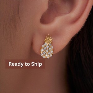 May include: Gold-colored pineapple stud earrings with clear, square-cut stones. The earrings are shaped like a pineapple, with a textured crown and a body covered in sparkling stones. The text "Ready to Ship" is displayed in a brown rectangle.