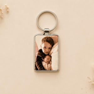 Peut inclure: Un porte-clés rectangulaire en métal argenté avec une insertion photo personnalisée. L'image montre un jeune enfant tenant un nouveau-né. Le porte-clés comprend un anneau fendu circulaire sur le dessus pour la fixation, avec un cadre métallique poli.