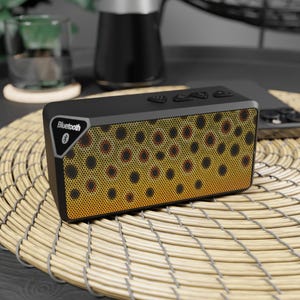 May include: A black rectangular Bluetooth speaker with a brown and yellow speckled front panel. The speaker has three buttons on top and the Bluetooth logo on the side. The speaker is sitting on a woven mat.