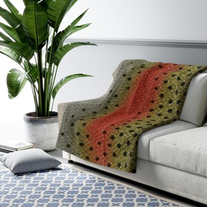 May include: A throw blanket draped over a white sofa, featuring a detailed print of a trout's skin. The design includes a central red stripe, surrounded by olive green and gray scales with black spots. A large potted plant is in the background.