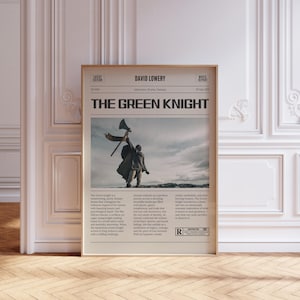 Op de afbeelding: Een ingelijste filmposter voor "The Green Knight" met een beige achtergrond. De poster toont een figuur met een bijl tegen een bewolkte lucht. Tekst bevat "David Lowery" en "Movie Report".