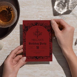 May include: A red birthday party invitation with black ornate detailing is held by hands. The invitation reads "Megan's Birthday Party 18". A glass of amber liquid with a star anise sits on a dark wooden tray.