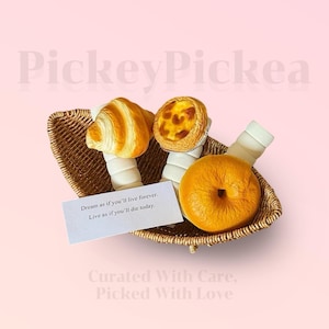 May include: A wicker basket containing a croissant, a custard tart, a bagel, and two wrapped items. A card reads "Dream as if you'll live forever. Live as if you'll die today." The background is a pale pink.