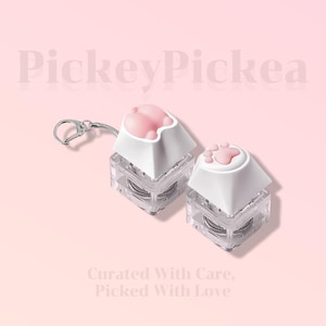 May include: Two keyboard keycaps with a cat paw design. The keycaps have a white base and a pink paw print on top. One keycap is attached to a silver keychain. The background is a light pink color with the text "PickeyPickea" and "Curated With Care, Picked With Love".
