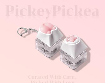 May include: Two keyboard keycaps with a cat paw design. The keycaps have a white base and a pink paw print on top. One keycap is attached to a silver keychain. The background is a light pink color with the text "PickeyPickea" and "Curated With Care, Picked With Love".