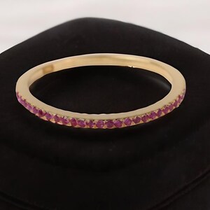 Peut inclure: Une bague en or ornée d'une rangée de petites pierres précieuses rouges. La bague est posée sur un écrin en velours noir, sur fond blanc. Un bijou délicat.