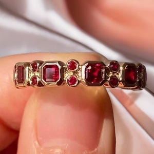 Peut inclure: Une bague en or ornée d'une série de pierres précieuses rouges de coupe carrée et ronde alternées. Les pierres sont serties dans une monture en or, créant un motif continu. La bague est présentée sur un fond blanc doux.