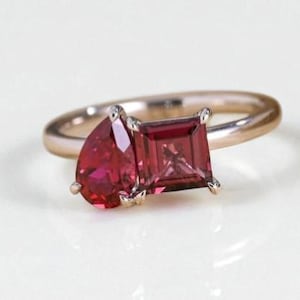 May include: A rose gold ring featuring two gemstones: a pear-shaped and a square-cut, both in a deep red hue. The ring's band is a simple, polished design, showcasing the vibrant stones.